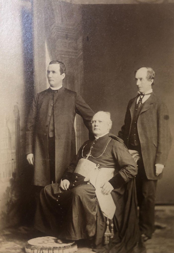 A historical photograph from the University Archives, in sepia tones. Pictured are Rev. Cornelius O'Brien, standing left; Bishop McIntyre of Charlottetown, sitting centre; and an unidentified layman, standing right.